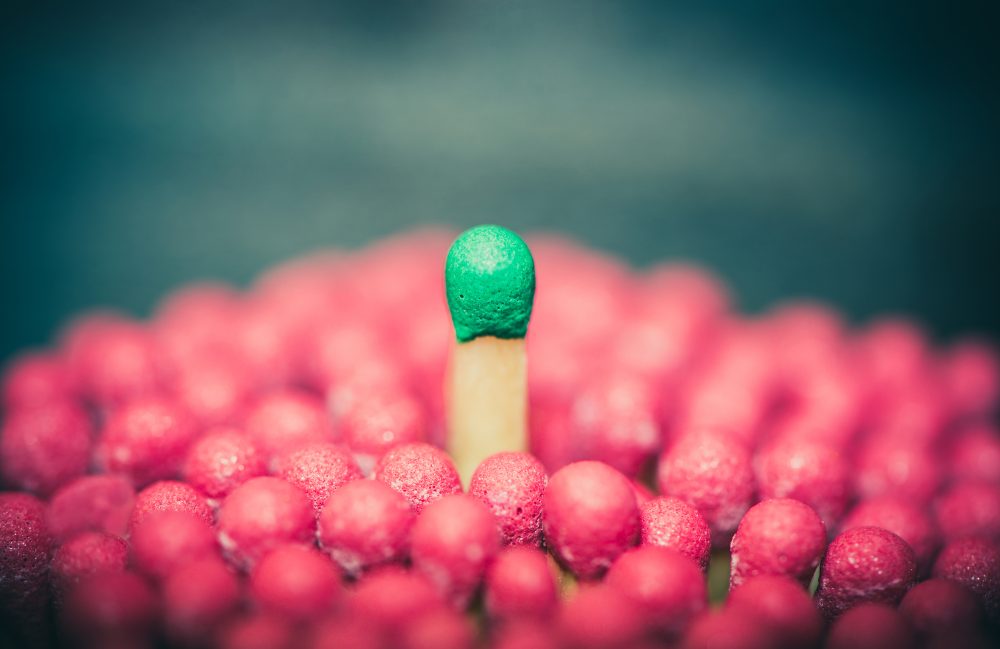 Image: wooden match with green top standing out from a field of wooden matches with red tops