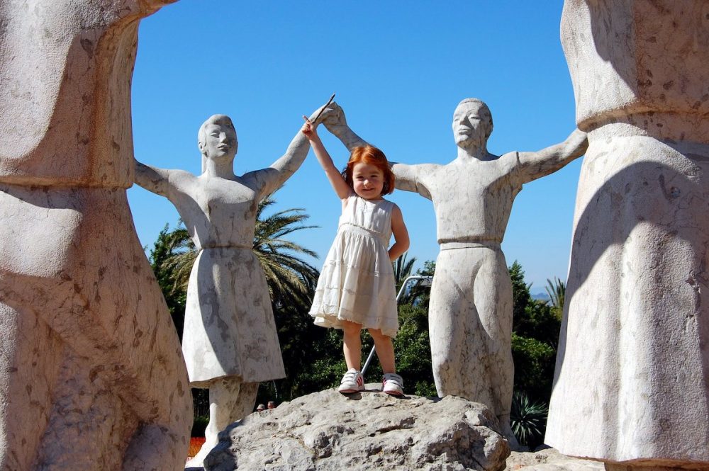 Image: young re-headed girl standing amid a ring of figurative statues