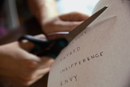 Image: person using scissors to cut a piece of paper on which are written the words "hatred," "indifference," and "envy"