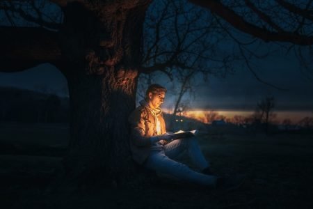 Image: young man sitting against a tree in the evening, reading a book from which emanates a glow