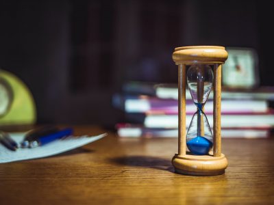 Image: small hourglass on a table with books, pens, and paper in the background.