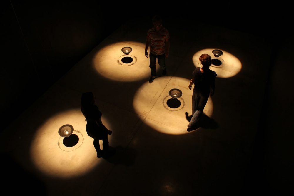 Image: art installation of a dark room containing four audio speakers suspended in mid-air and lit by spotlights, with people walking through the space.