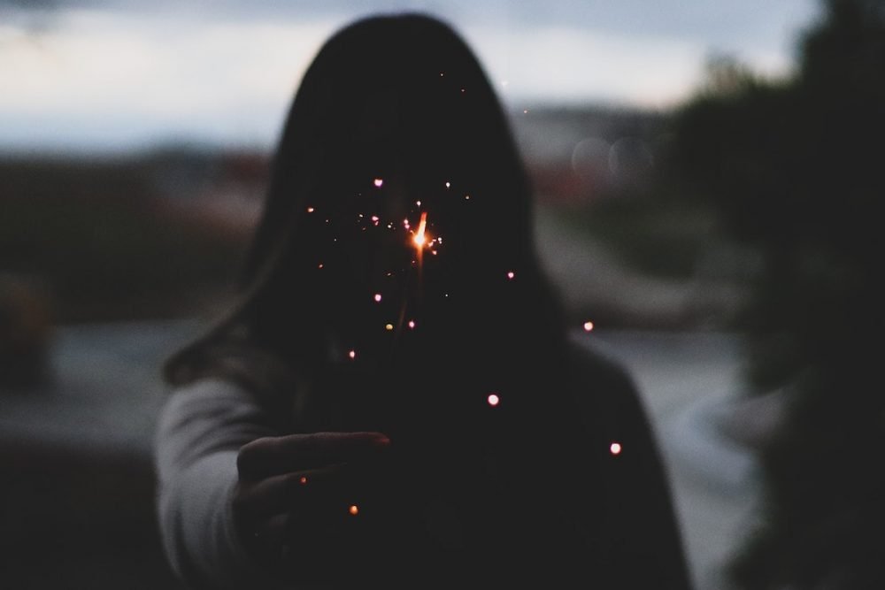 Image: a woman in silhouette holding forth a lit sparkler.