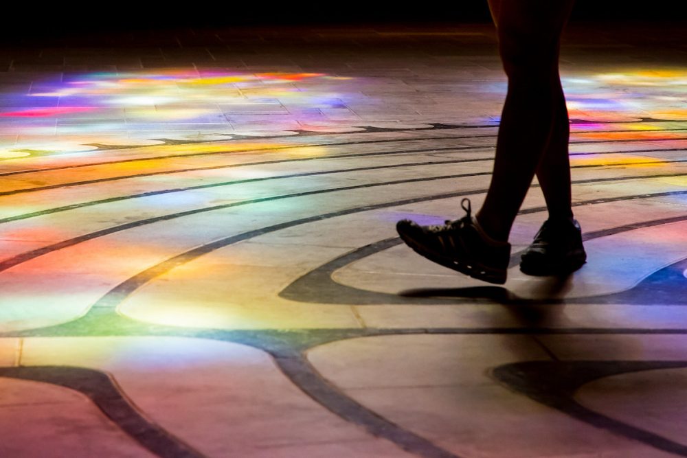 Image: a person walking on a floor which is decorated with curved patterns and illuminated with light from a multi-colored window.