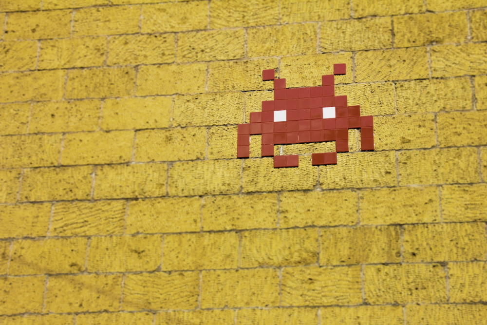Image: street art of a pixelated video-game style space invader made of red tiles applied to a yellow brick wall.