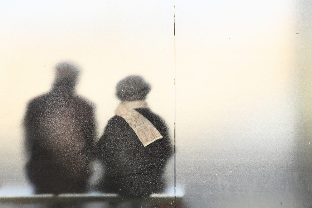 Image: silhouettes of an older couple sitting at a bus stop, seen from the back through frosted glass.