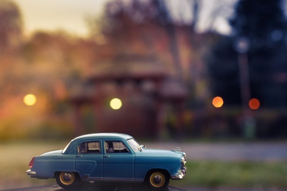 Image: close-up photo of a miniature antique car in the foreground, with blurred city street in the background.