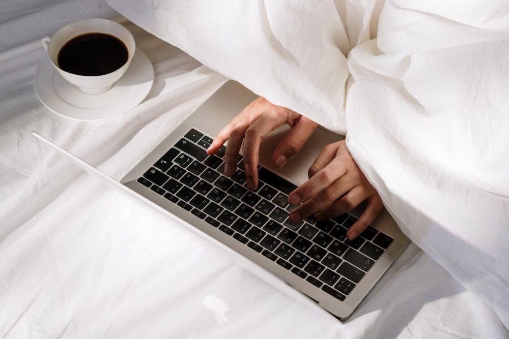 Image: hands emerging from bed covers to type on a laptop computer, with a cup of coffee resting nearby on the bedsheets.