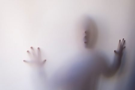 Image: an indistinct figure's hands and face behind a white screen.