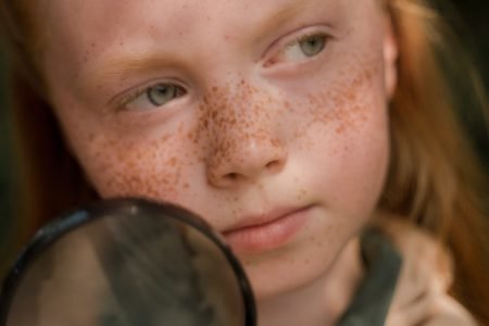 Image: a young, redheaded, freckled girl holding a magnifying glass.