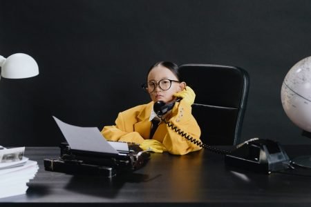 Image: young girl wearing an adult-sized suit, sitting at an office desk and holding a telephone receiver.