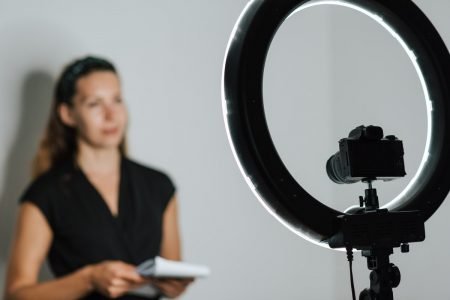 Image: a woman holding a notepad, standing in front of a camera and ring light mounted on a tripod.