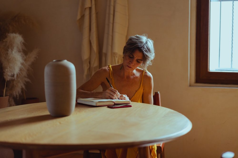 Image: a woman alone at a table, writing in a journal.