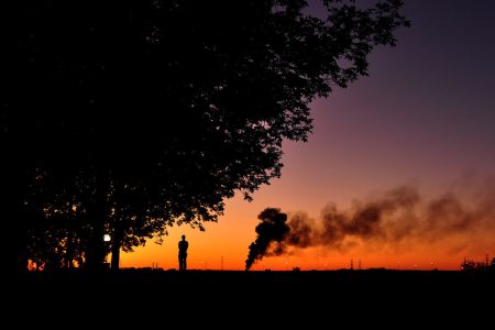 Image: a lone person stands watching distant smoke rising, silhouetted against a colorful sunrise.