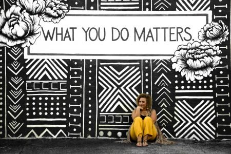 Image: a woman sitting against a wall on which is painted a black and white mural and the words "What you do matters."
