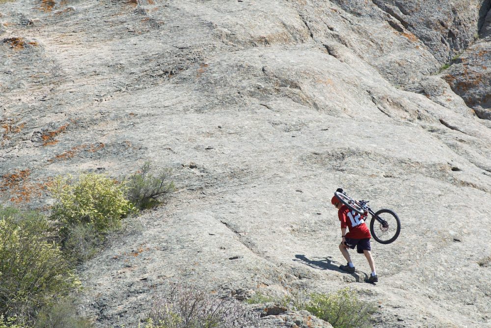 Image: a mountain biker climbing a mountain on foot, carrying his bicycle on his back.