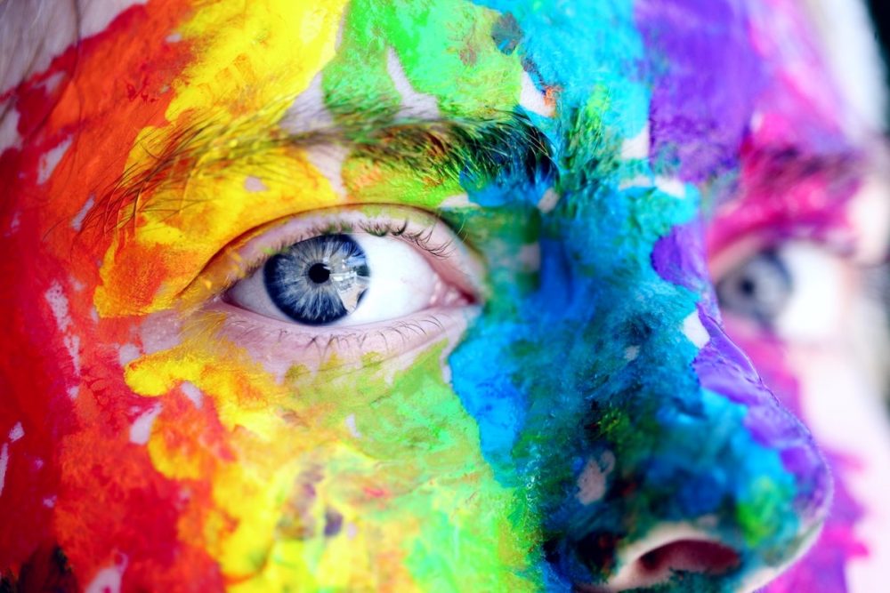 Image: close-up photo of a face painted with rainbow colors, the subject's eyes gazing intently at the viewer.