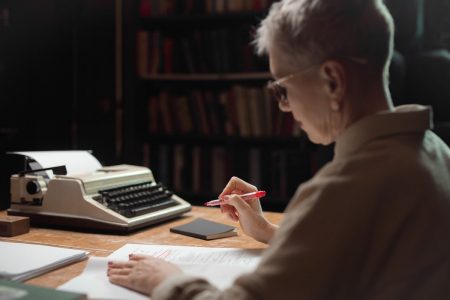 Image: a woman sitting at a desk, editing a manuscript with a red pen.