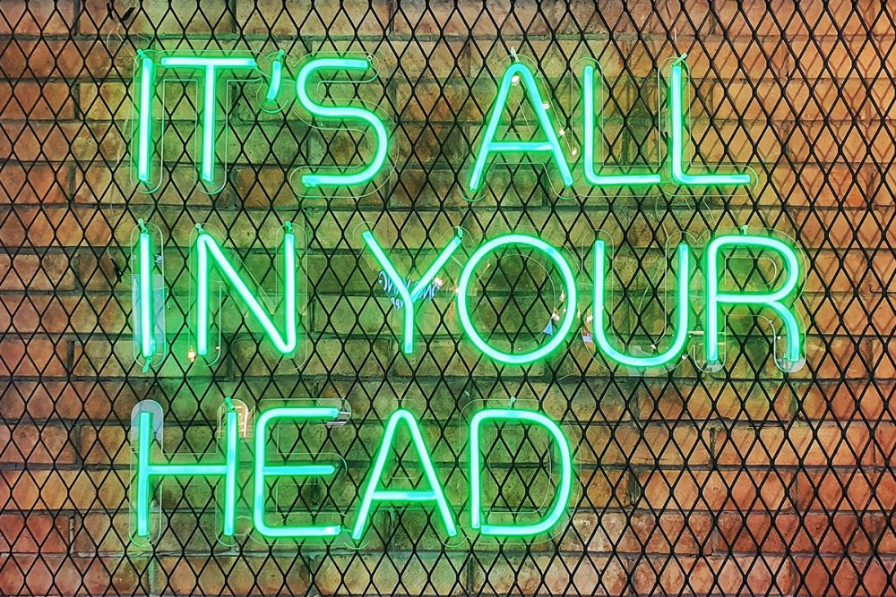 Image: a green neon sign reading "It's all in your head" is mounted on a metal mesh frame against a brick wall.