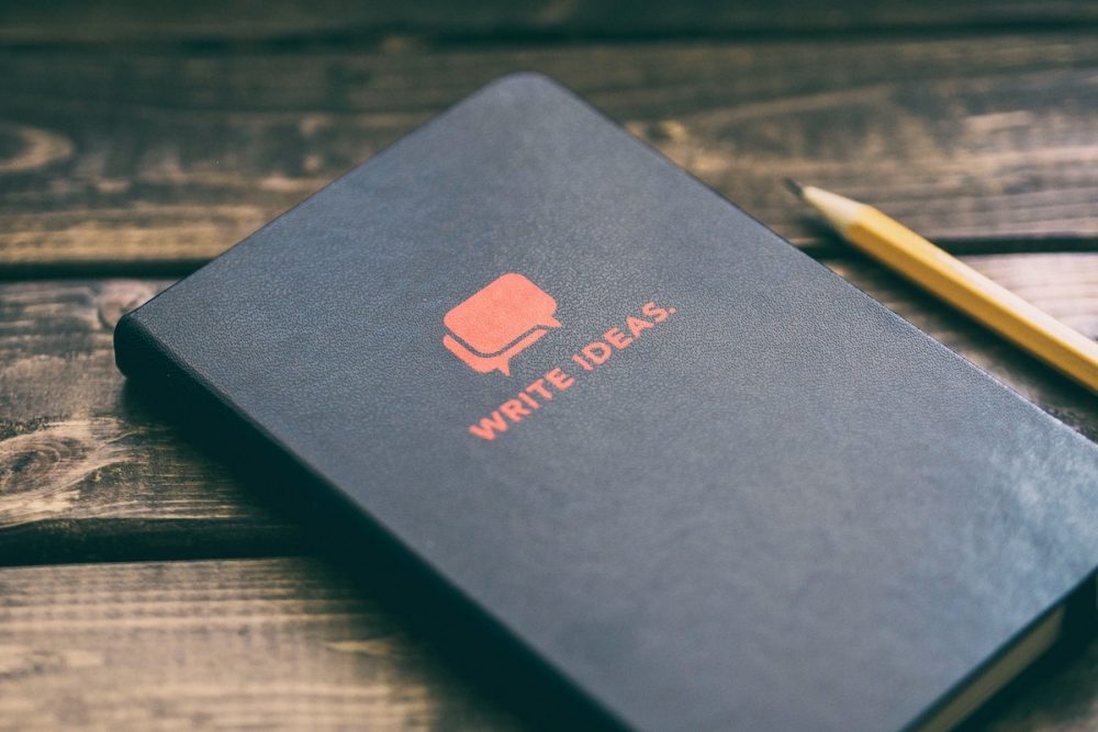 Image: a closed notebook emblazoned with a logo that reads "Write ideas" sits on a wooden table alongside a pencil.