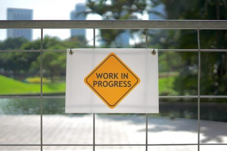 Image: a yellow and white sign reading "work in progress" attached to a wire fence.