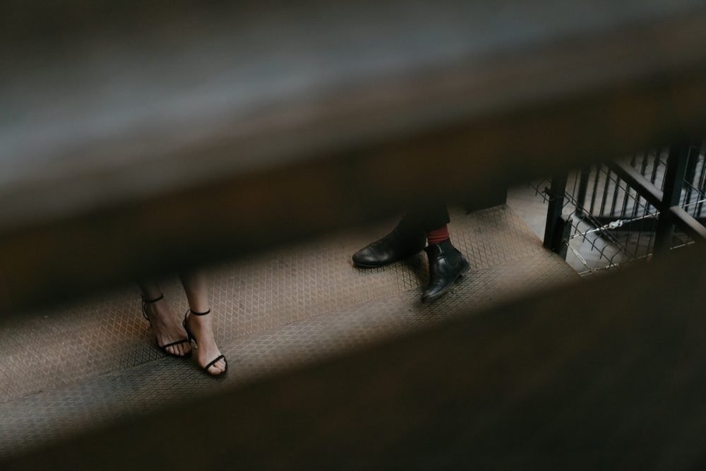 Image: through a gap between stairs are visible just the feet of a woman and a man on a train platform.