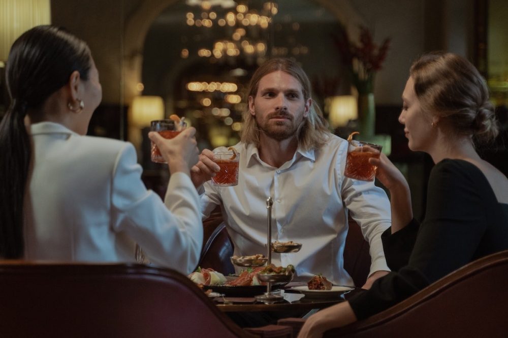 Image: a man and two women sit with cocktails in hand around a restaurant table.