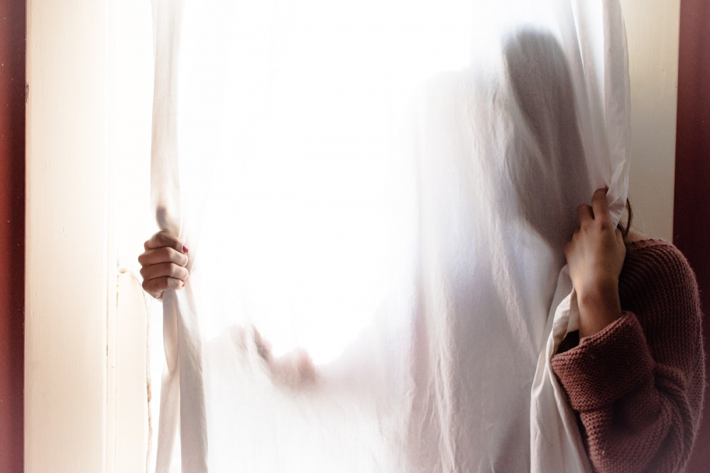 Image: a woman hides behind a curtain, only her hands and shoulder visible.