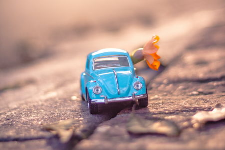 Image: close-up photo of a miniature, blue diecast Volkswagen Beetle with a flower protruding from its window, placed on a cobblestone street.