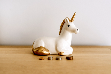 Image: small piles of coins rest on a table in front of a porcelain bank shaped like a unicorn.