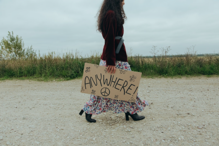 Image: Woman hitchhiker in bohemian clothing, walking along a dirt road and holding a handmade cardboard sign reading "Anywhere!"