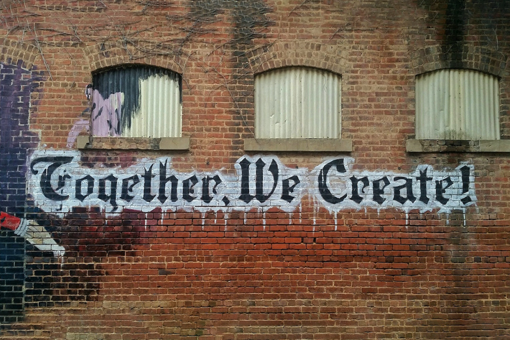Image: exterior brick wall of an abandoned factory, on which is painted a mural reading "Together, We Create!"
