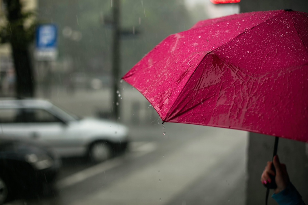 Image: a woman's hand holds a magenta umbrella as she shelters from rain in an urban doorway.