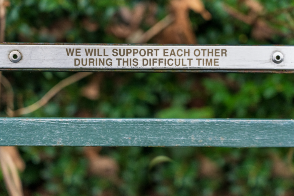 Image: one of the slats at the back of a park bench bears a plaque reading "We will support each other during this difficult time."