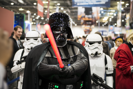 Image: at a comicon, a Darth Vader cosplayer points a lightsaber at the viewer while two Stormtroopers stand behind.