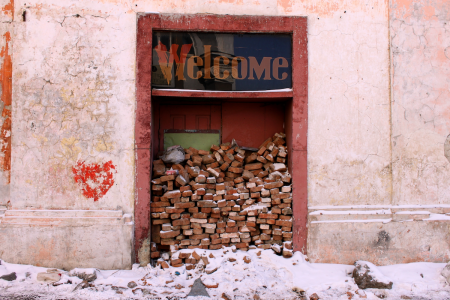 Image: the doorway of a decrepit building is piled with bricks, preventing entrance. Above the doorway is a large sign saying "Welcome."
