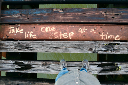 Image: a person stands on a rotting wooden plank of a derelict footbridge. Between planks are large gaps through which water is visible. On one plank are painted the words "Take life one step at a time."
