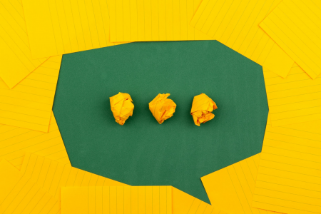 Image: yellow notepad paper is arranged on a green background to represent the shape of a cartoon speech bubble, with three crumpled wads of yellow paper aligned like an ellipsis in the center.