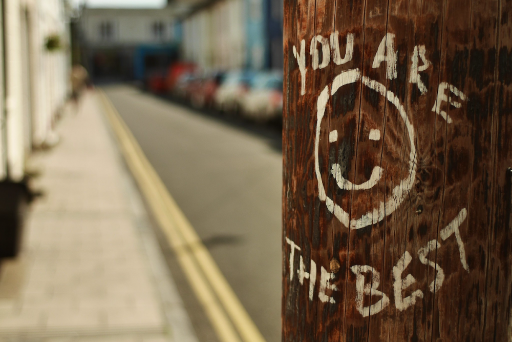 Image: an urban telephone pole on which someone has painted a smiley face and the words "You are the best".
