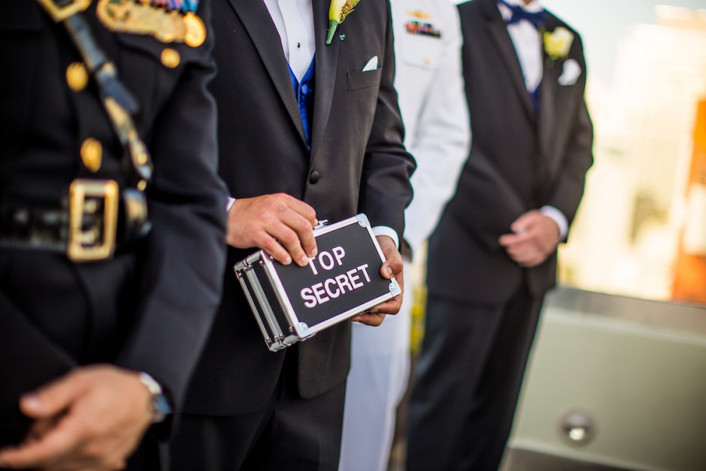 Image: in a row of men garbed in formal evening wear and military dress, one of them holds a small box labeled "Top Secret."