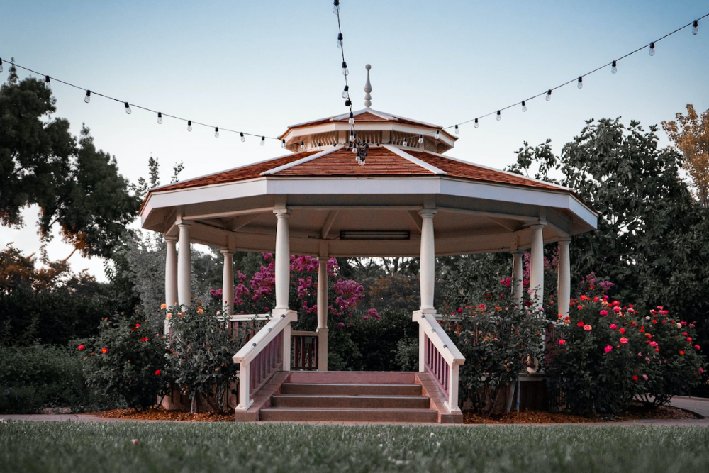 From an ant's-eye view, a pretty gazebo is surrounded by blooming rose bushes and festooned with lights on a clear summer day.