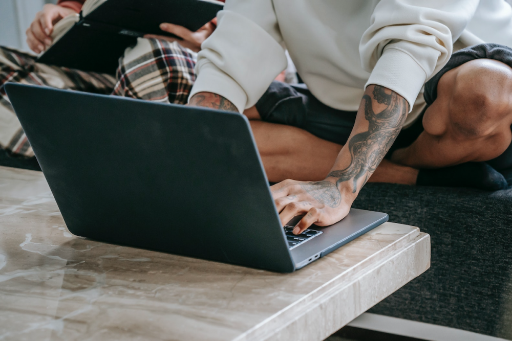 Image: a heavily-tattooed man in casual clothing sits cross-legged on a sofa and types at a laptop computer, while someone else sits beside him reading a book.