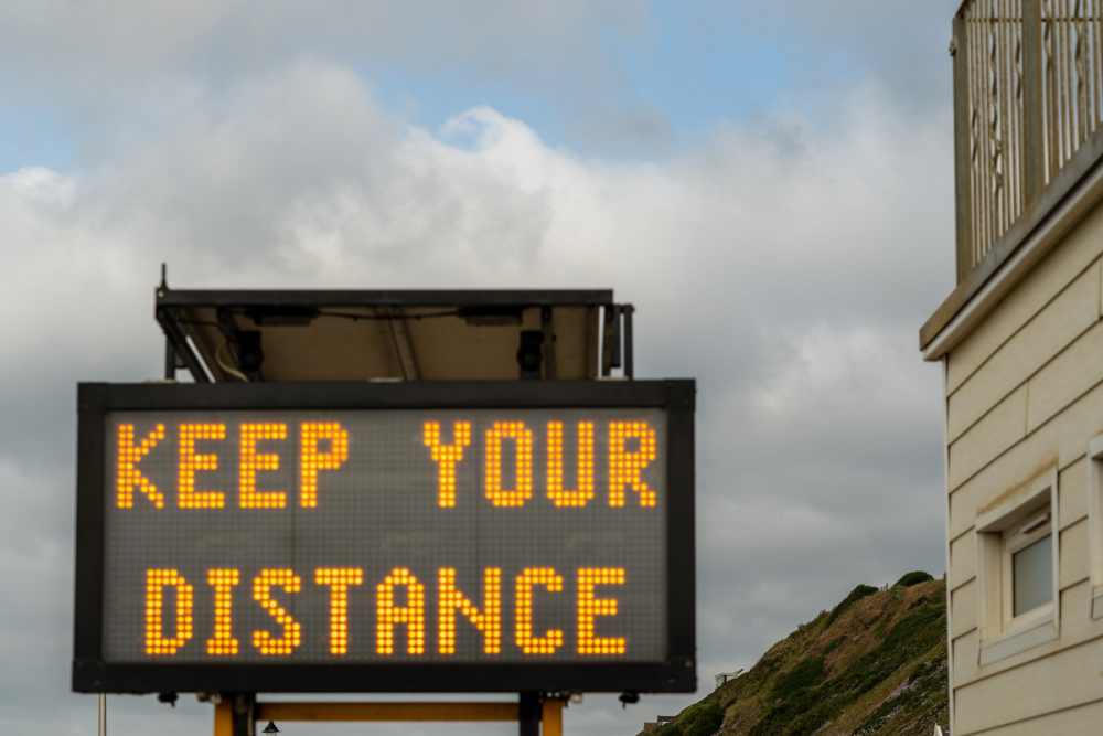 Image: next to a roadway, a lighted traffic control sign displays the message "Keep Your Distance."