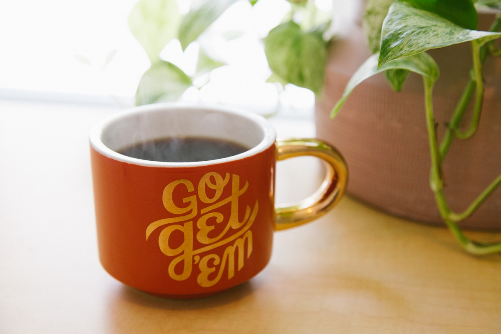 Image: next to a potted plant and sunny window is a mug of steaming coffee emblazoned with the words "Go get 'em."