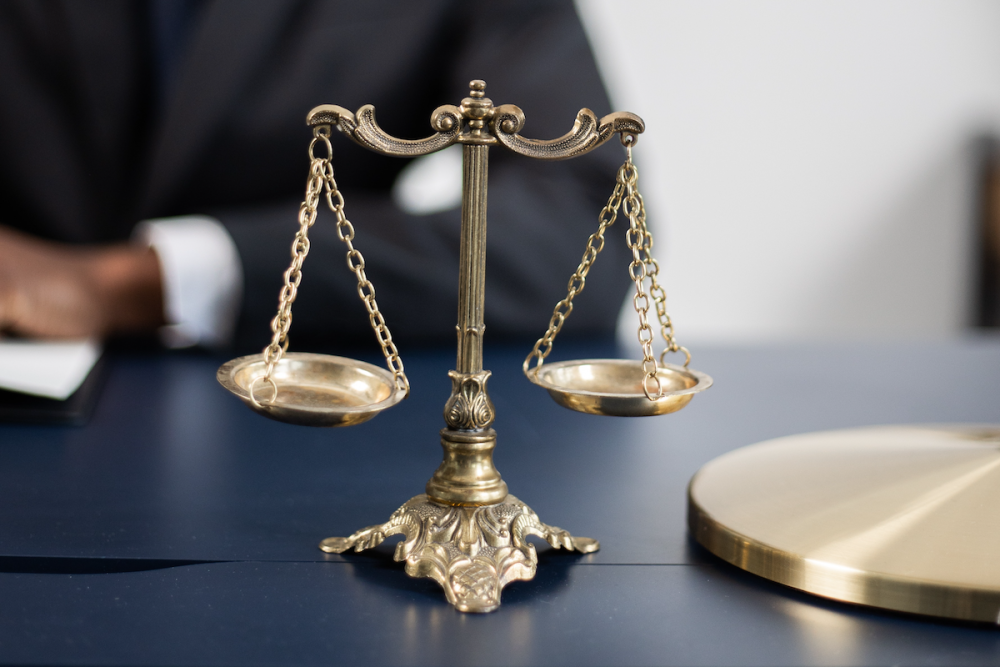 Miniature gold-plated scales of justice sit on an office desk in front of a man wearing a business suit.