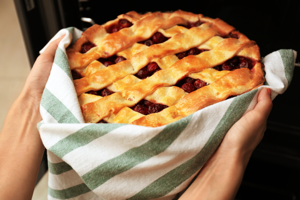 A woman's hands cradle a freshly baked cherry pie in a tea towel as she removes it from the oven.