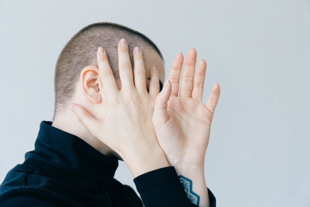 A woman with closely-cropped hair uses her hands to hide her face from the viewer.