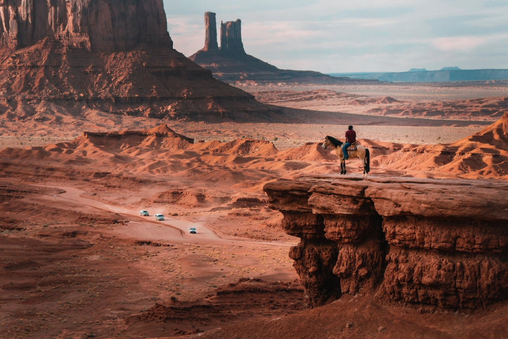 A Native American man wearing street clothes sits astride a horse atop a bluff overlooking the landscape of Oljato Monument Valley in Arizona. On a road below, two vans and a car drive along a winding road.