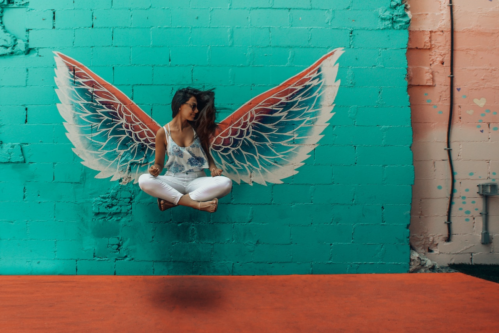 Image: a colorful pair of wings are painted on a cinderblock wall. Seeming to float midair in front of the wings as if they belong to her, a woman sits crosslegged.