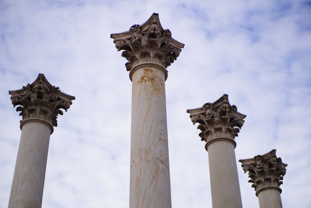 Image: Four white corinthian columns stand against a partly-cloudy blue sky.